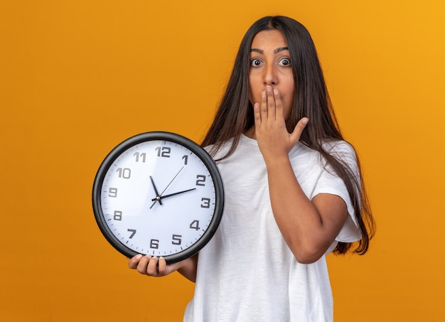 Young girl in white t-shirt holding wall clock looking at camera being shocked covering mouth