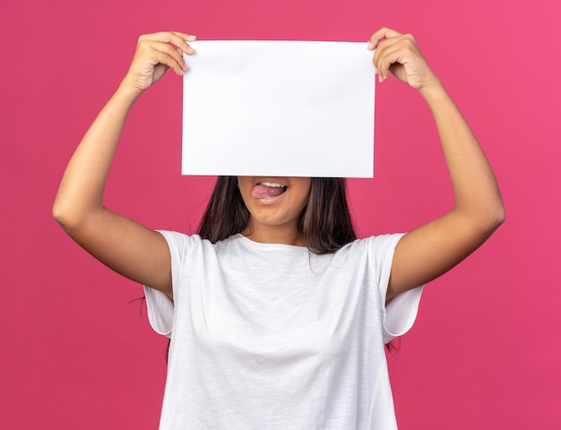 Young girl in white t-shirt holding white blank sheet of paper in front of her face smiling standing over pink