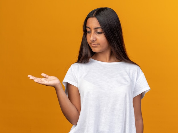 Young girl in white t-shirt looking aside with confident expression presenting copy space with arm of her hand standing over orange background