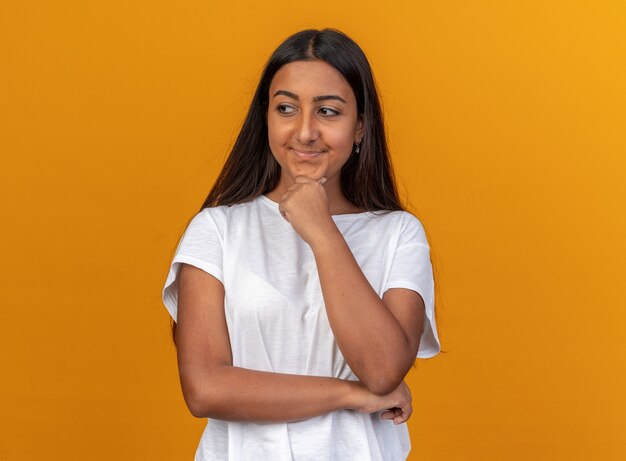 Young girl in white t-shirt looking aside with hand on her chin smiling standing over orange background