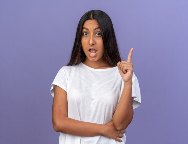 Young girl in white t-shirt looking at camera amazed and surprised showing index finger having new idea standing over blue background