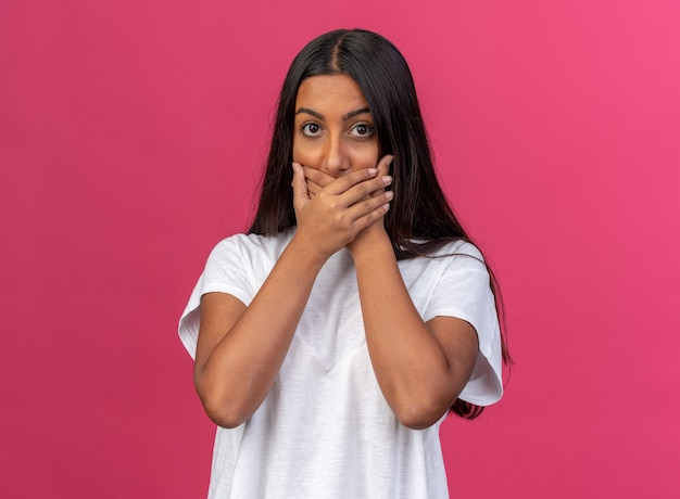 Young girl in white t-shirt looking at camera being shocked covering mouth with hands standing over pink background