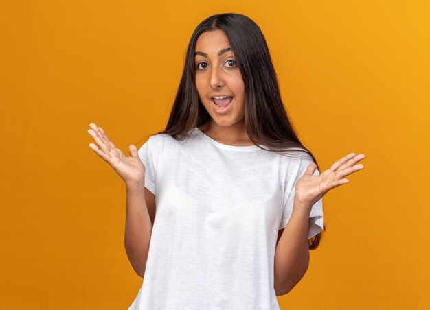 Young girl in white t-shirt looking at camera happy and cheerful smiling raising arms standing over orange