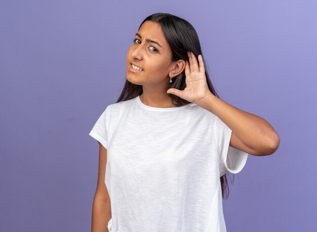 Young girl in white t-shirt looking at camera intrigued with hand over her ear trying to listent to something standing over blue background