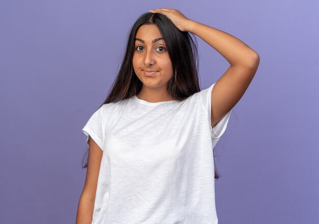 Young girl in white t-shirt looking at camera smiling being confused with hand on her head for mistake 