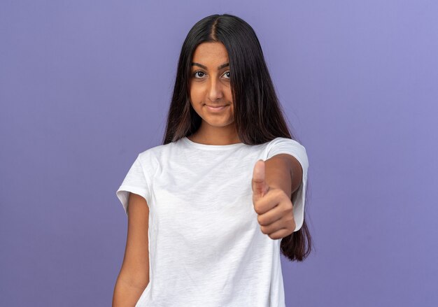 Young girl in white t-shirt looking at camera smiling confident showing thumbs up standing over blue