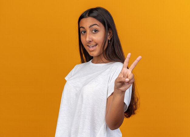Young girl in white t-shirt looking at camera smiling confident showing v-sign standing over orange
