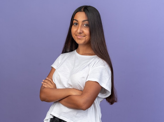 Foto gratuita giovane ragazza in maglietta bianca che guarda l'obbiettivo sorridente sicura di sé con le braccia incrociate sul petto in piedi sopra il blu