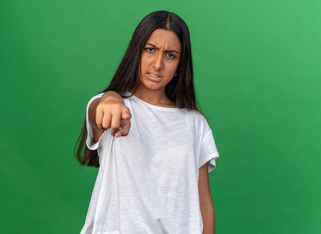 Young girl in white t-shirt looking at camera with angry face pointing with index finger at camera standing over green background