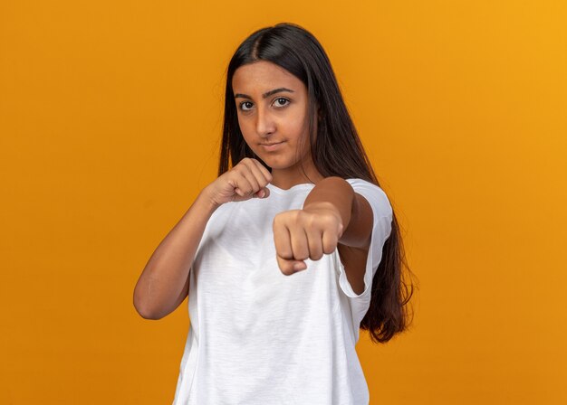 Молодая девушка в белой футболке смотрит в камеру со сжатыми кулаками, позирует как боксер с серьезным лицом