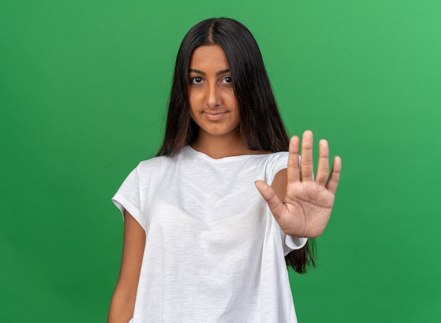 Young girl in white t-shirt looking at camera with confident smile on face making stop gesture with hand 