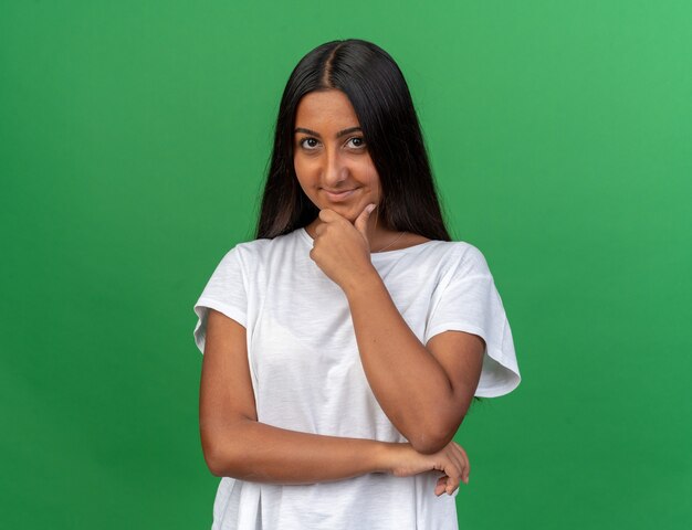 Young girl in white t-shirt looking at camera with hand on chin smiling standing over green background