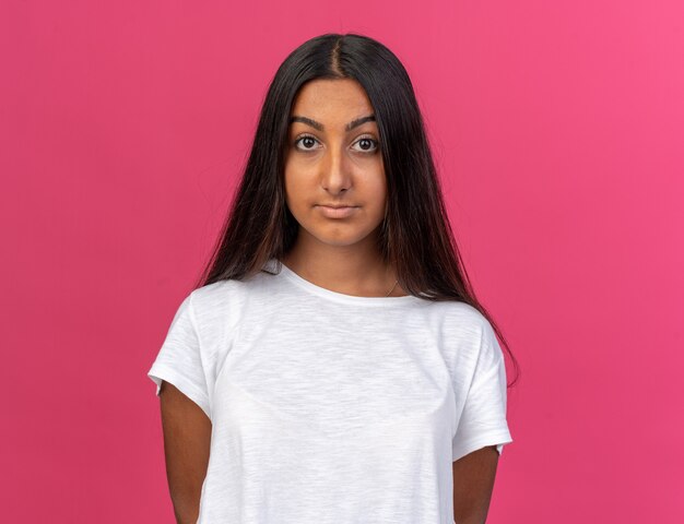 Young girl in white t-shirt looking at camera with serious confident expression standing over pink