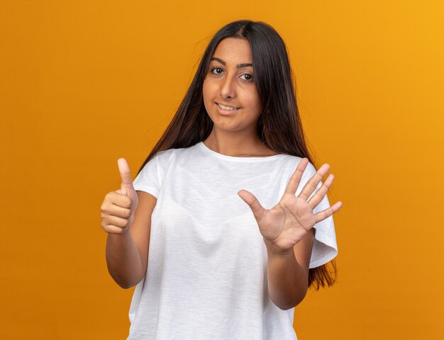 Young girl in white t-shirt looking at camera with smile on face showing and pointing up with fingers number six 