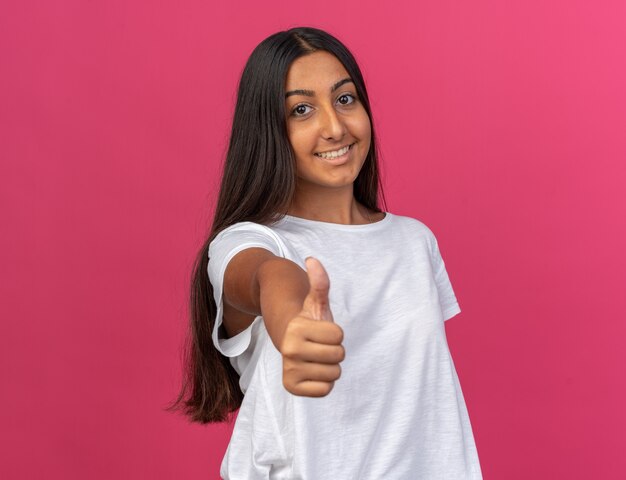 ピンクの上に立って親指を示す幸せそうな顔に笑顔でカメラを見て白いTシャツの少女