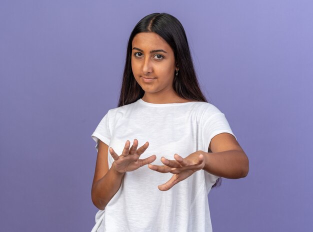 Young girl in white t-shirt looking at camera worried making defense gesture with hands standing over blue