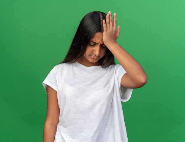 Young girl in white t-shirt looking confused and very anxious with hand on her head fro mistake standing over green background