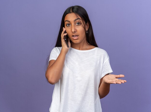 Young girl in white t-shirt looking confused while talking on mobile phone standing over blue background