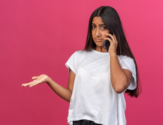 Young girl in white t-shirt looking confused while talking on mobile phone with raised arm standing over pink background