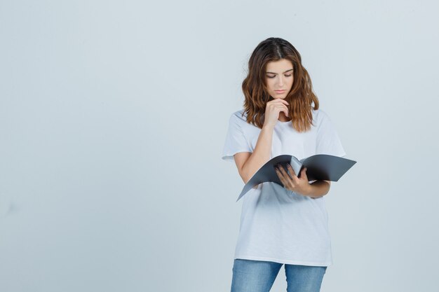 Молодая девушка в белой футболке просматривает заметки в папке и нерешительно смотрит, вид спереди.