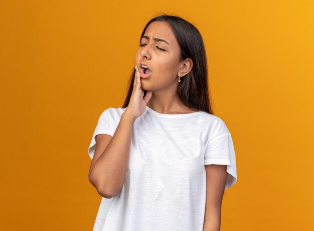 Young girl in white t-shirt looking unwell touching her cheek feeling toothache standing over orange