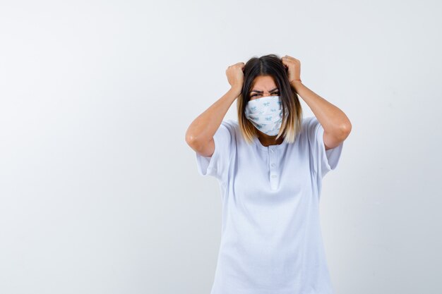 Young girl in white t-shirt and mask holding hands on head and looking annoyed , front view.