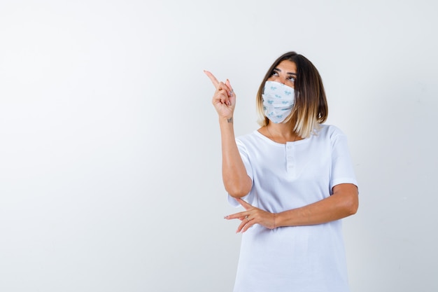 Young girl in white t-shirt and mask holding one hand under elbow, pointing up with index finger and looking focused , front view.