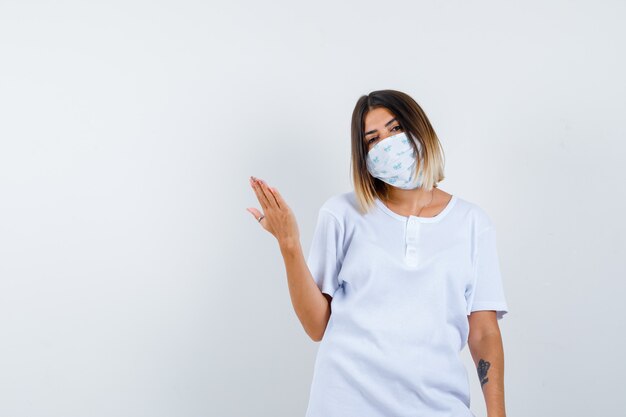 Young girl in white t-shirt and mask holding palm out and looking confident , front view.