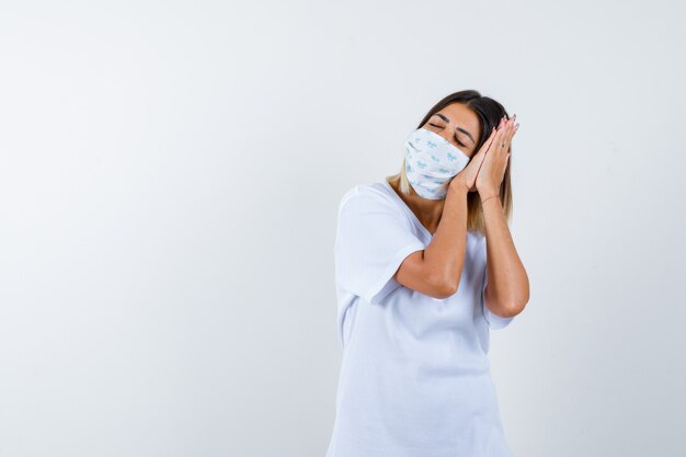 Young girl in white t-shirt and mask leaning cheek on palms as pillow and looking sleepy , front view.