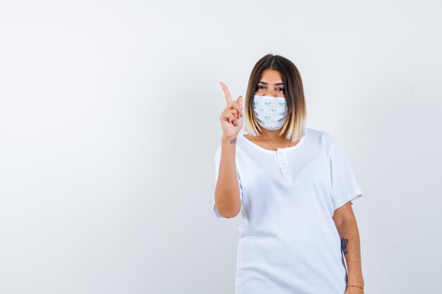 Young girl in white t-shirt and mask raising index finger in eureka gesture and looking sensible , front view.