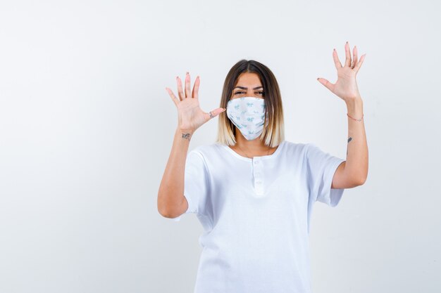 Young girl in white t-shirt and mask raising palms in surrender gesture and looking happy , front view.