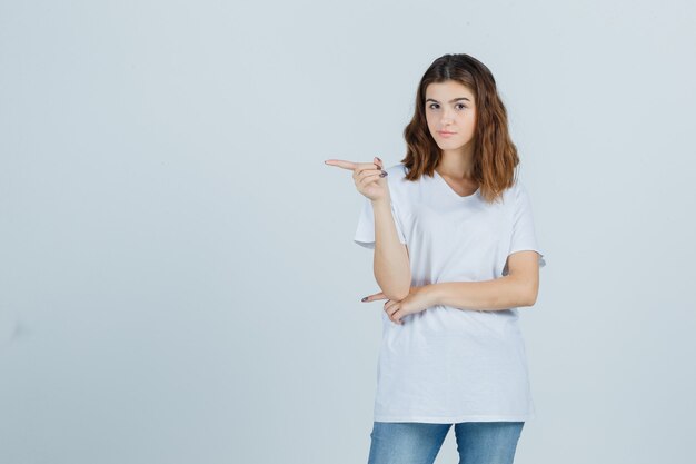 Young girl in white t-shirt pointing to the left side and looking pensive , front view.