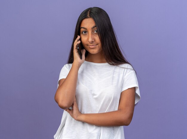 Молодая девушка в белой футболке дружелюбно улыбается, глядя в камеру во время разговора по мобильному телефону, стоя на синем фоне