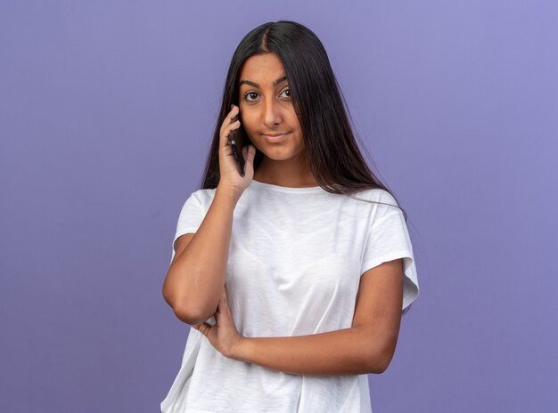 Young girl in white t-shirt smiling friendly looking at camera while talking on mobile phone standing over blue background