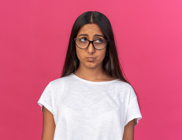 Young girl in white t-shirt wearing glasses looking aside with sad expression pursing lips standing over pink