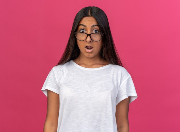 Young girl in white t-shirt wearing glasses looking at camera amazed and surprised standing over pink