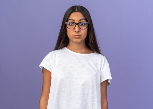 Молодая девушка в белой футболке в очках смотрит в камеру с серьезным уверенным выражением лица, стоящим над синим