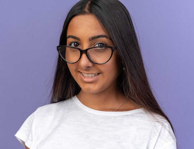 Young girl in white t-shirt wearing glasses looking at camera with smile on happy face standing over blue