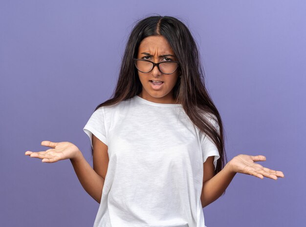 Young girl in white t-shirt wearing glasses looking confused and displeased raising arms in displeasure