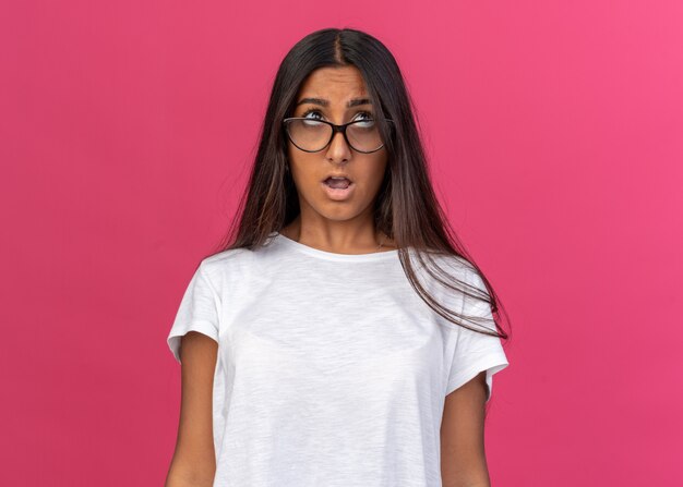 Young girl in white t-shirt wearing glasses looking up confused standing over pink background