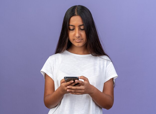 파란색 위에 서있는 스마트 폰을 사용하여 문자 메시지를 작성하는 흰색 티셔츠에 어린 소녀
