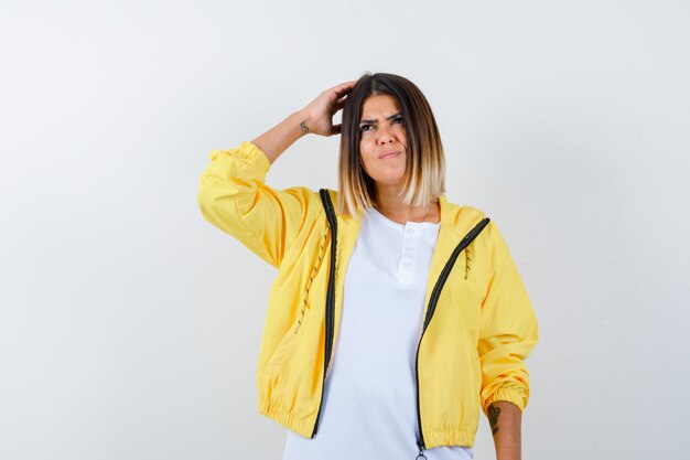 Young girl in white t-shirt , yellow jacket scratching head, thinking about something and looking pensive , front view.