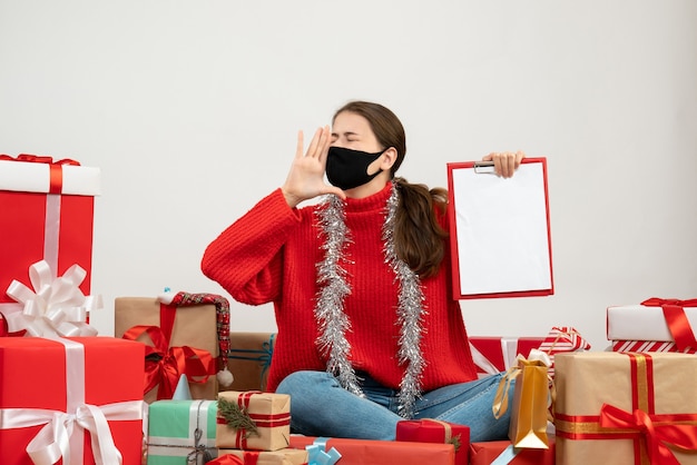 young girl with black mask holding documents and shouting sitting around presents on white