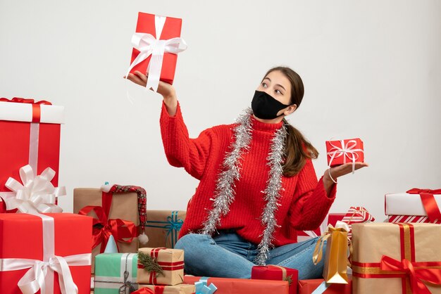 young girl with black mask holding gifts in both hands sitting around presents on white