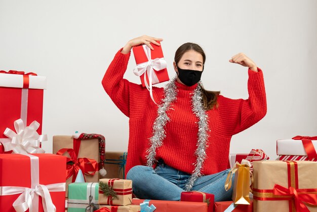 young girl with black mask holding present showing her muscle sitting around presents on white