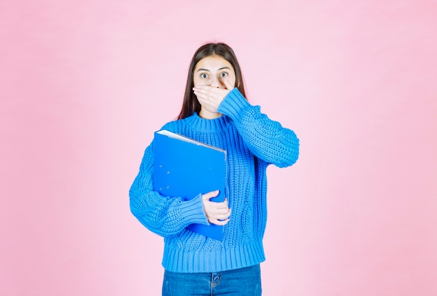 Free photo young girl with a blue folder coning mouth with hand on pink.