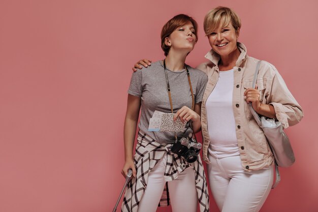 Young girl with brunette short hair blowing kiss. holding tickets and camera and posing with blonde lady in white outfit on pink backdrop. 