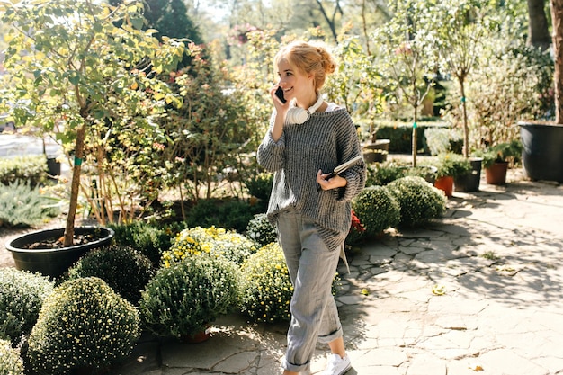 無料写真 お団子を持つ少女は植物園の路地を歩いて電話で話している彼女の首にヘッドフォンと彼女の手にノートを持ったきれいな女性の肖像画