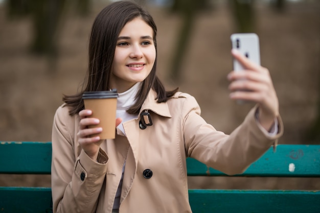 Young girl with coffee cup makes selfie in the autumn park