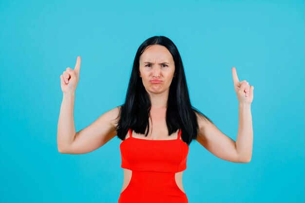 Young girl with dissatifies mimicry is pointing up with forefingers on blue background